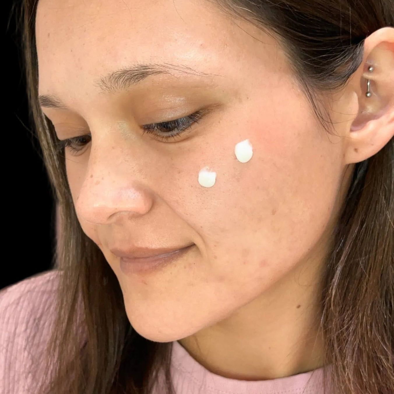Woman with two dots of Luminous Empire Collagen cream on her face, wearing a pink sweater.